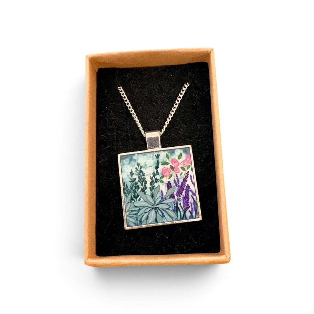 A square pendant necklace with a colorful floral and botanical illustration, displayed inside an open tan gift box with a black velvet lining on a white background.
