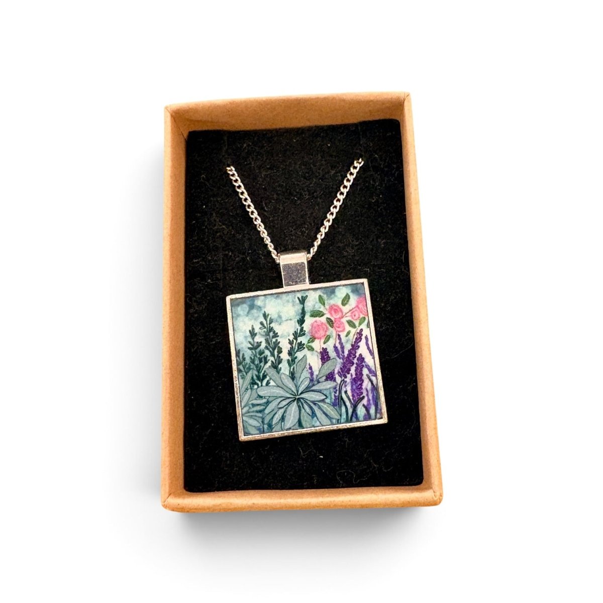 A square pendant necklace with a colorful floral and botanical illustration, displayed inside an open tan gift box with a black velvet lining on a white background.