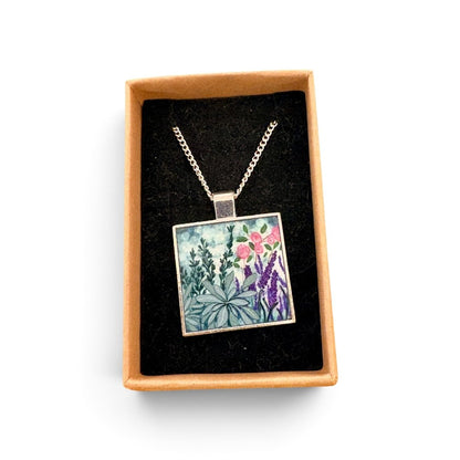 A square pendant necklace with a colorful floral and botanical illustration, displayed inside an open tan gift box with a black velvet lining on a white background.