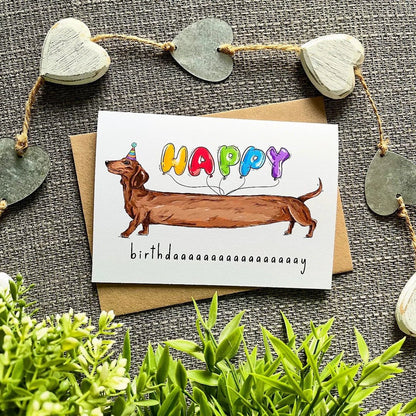 Birthday card with a dachshund and colorful balloons on a textured surface with heart decorations.