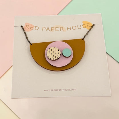 A handmade geometric pendant necklace from Red Paper House, featuring a mustard yellow semicircle with a pink and light blue circular center containing patterned and solid details, displayed on its branded white card.