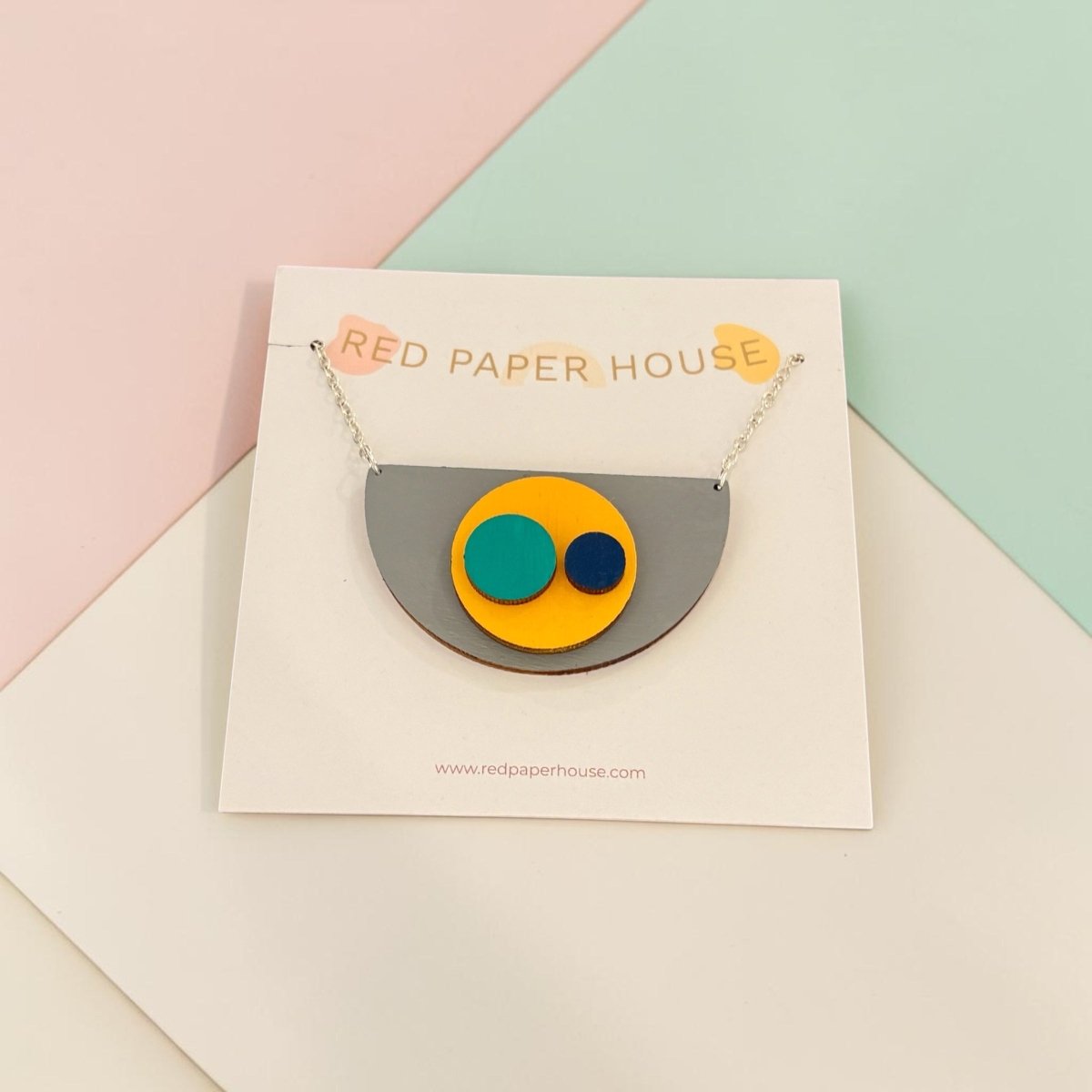 A geometric acrylic pendant necklace from Red Paper House, featuring a gray semicircle with yellow, teal, and navy circles, displayed on a white branded card against a pastel pink, green, and white background.