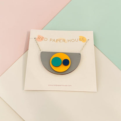 A geometric acrylic pendant necklace from Red Paper House, featuring a gray semicircle with yellow, teal, and navy circles, displayed on a white branded card against a pastel pink, green, and white background.