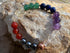 A multicolor chakra bead stretch bracelet arranged in a circle on a textured beige rock. The beads represent the seven chakras in color order: red, orange, dark blue, black, copper, clear, purple, white, and green.