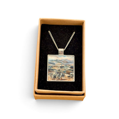 A square pendant necklace displayed in an open tan gift box with a black velvet insert. The pendant features a colorful landscape painting with blue, green, and beige tones, suspended from a silver-toned chain.