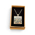 A square pendant necklace displayed in an open tan gift box with a black velvet insert. The pendant features a colorful landscape painting with blue, green, and beige tones, suspended from a silver-toned chain.