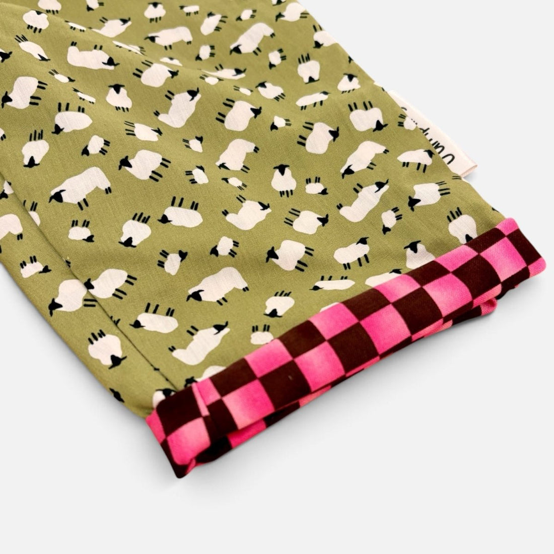 A folded piece of fabric with an olive green background printed with white sheep and black faces, finished with a folded over edge showing a hot pink and dark red checkered pattern.