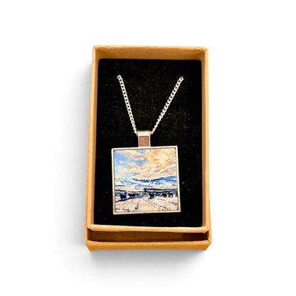 A square pendant necklace displayed in an open tan gift box with a black velvet insert. The pendant features a colorful landscape painting with blue, white, and yellow tones, suspended from a silver-toned chain.