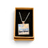 A square pendant necklace displayed in an open tan gift box with a black velvet insert. The pendant features a colorful landscape painting with blue, white, and yellow tones, suspended from a silver-toned chain.
