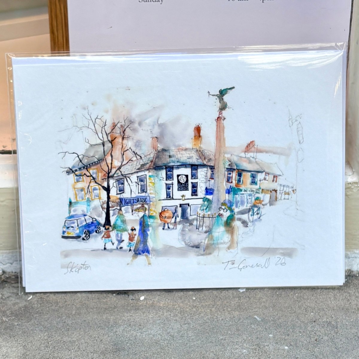 A watercolor art print depicting a street scene with buildings, pedestrians, and a tree, shown in a clear plastic sleeve on a gray floor. The print has a white border and handwritten signatures at the bottom.