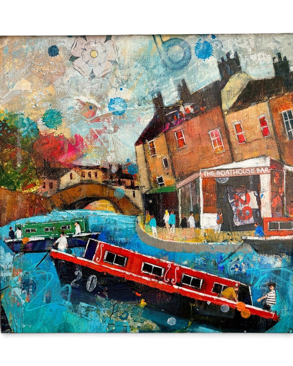 An expressive, colorful painting of a canal-side town scene with narrowboats on blue water, brick buildings, a bridge, and a pub, shown in a light wood frame.