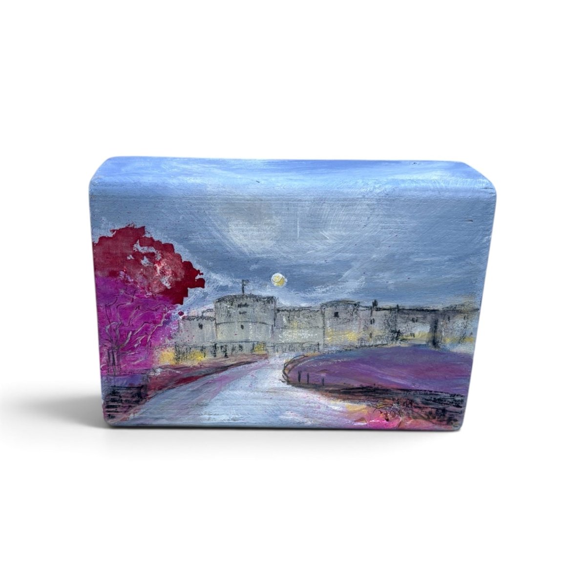 A rectangular wooden block painted with a landscape scene featuring a castle, a winding road, purple fields, and a red tree under a blue sky.