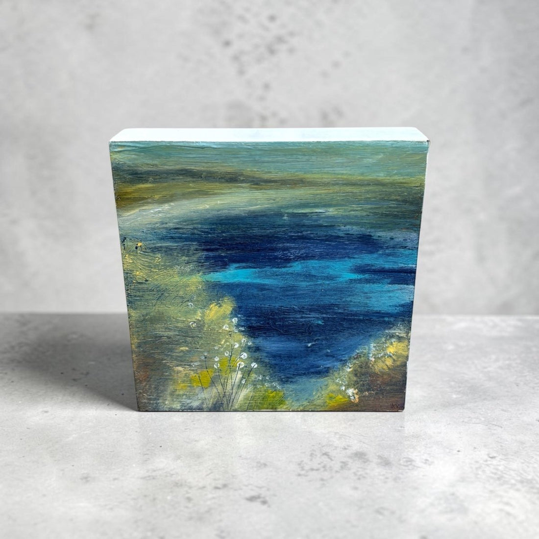 A small rectangular canvas painting of an abstract landscape with deep blue water, green and yellow foliage, and a pale sky, displayed on a light gray surface against a textured gray wall.