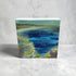 A small rectangular canvas painting of an abstract landscape with deep blue water, green and yellow foliage, and a pale sky, displayed on a light gray surface against a textured gray wall.
