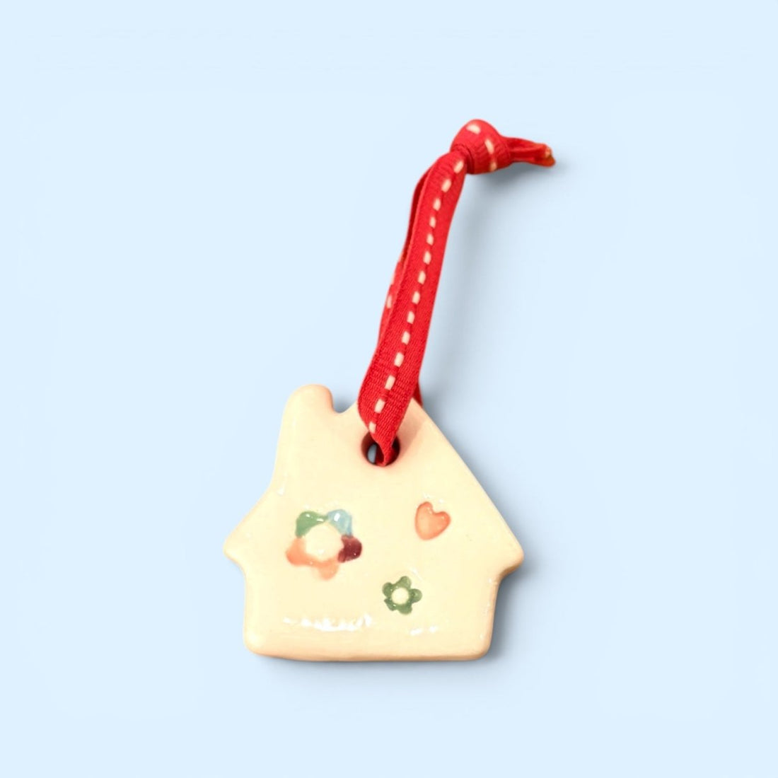A small house-shaped ceramic ornament in a warm cream color, decorated with colorful painted flowers and a red heart, hanging from a red ribbon with white dots, set against a solid light blue background.