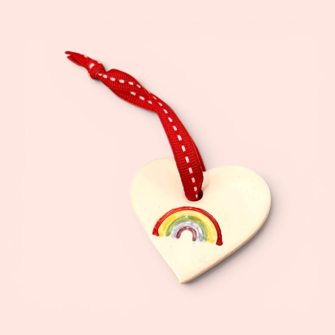 A heart-shaped wooden ornament painted with a small rainbow, hanging from a red ribbon with white stitching, set against a solid pale pink background.