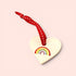 A heart-shaped wooden ornament painted with a small rainbow, hanging from a red ribbon with white stitching, set against a solid pale pink background.