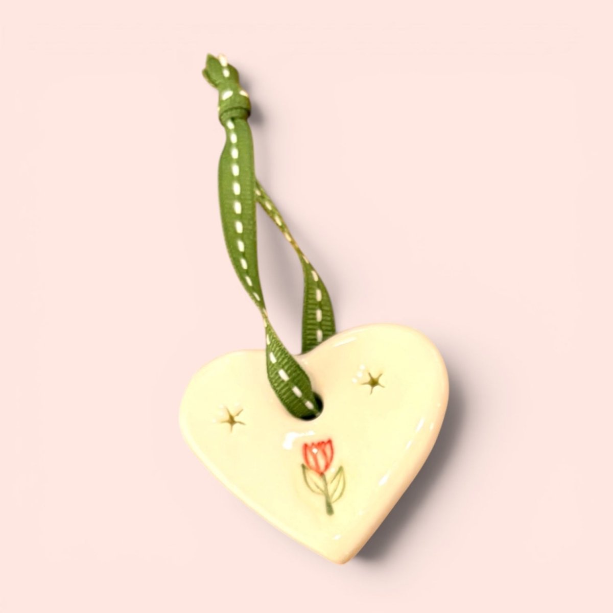 A glossy cream-colored ceramic heart ornament with a green and white polka-dot ribbon loop, decorated with a small red tulip and gold stars on a pale pink background.