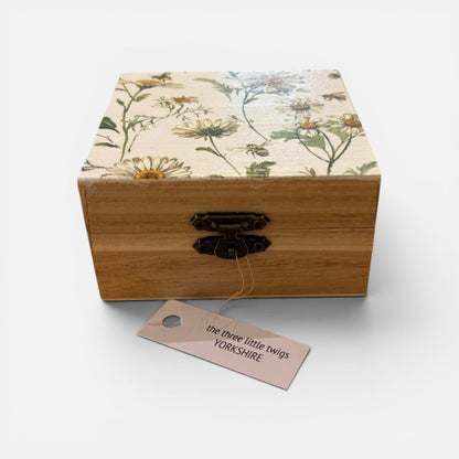 A square wooden keepsake box with a floral-patterned lid and a dark metal clasp, shown on a light gray background with a hanging tag that reads &