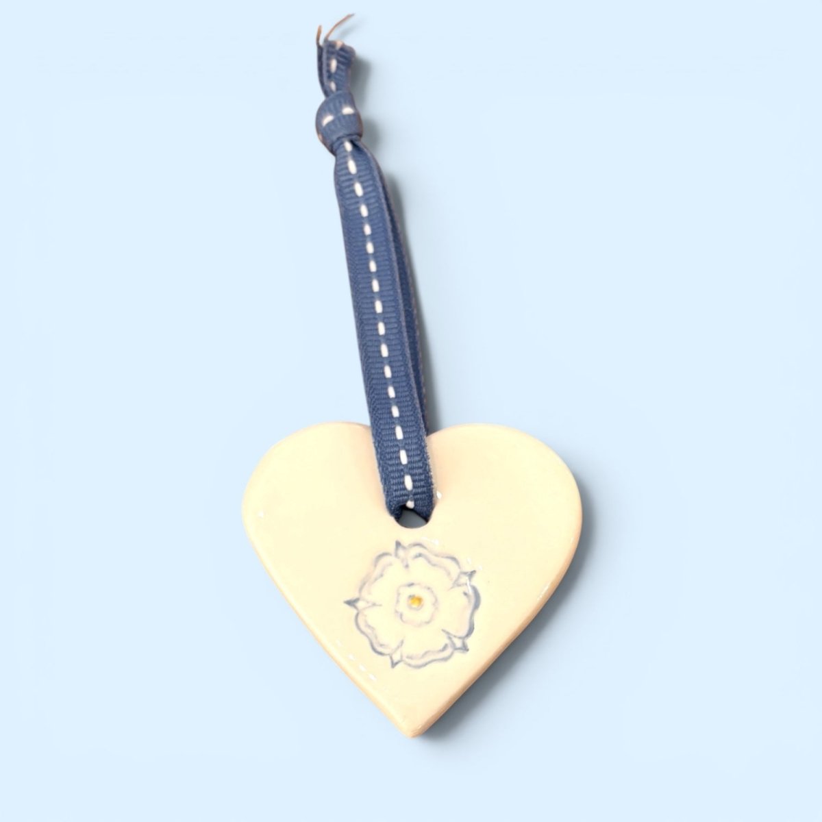 A heart-shaped wooden ornament in a pale cream color, featuring a hand-painted rose design and a navy blue ribbon hanger with white stitching, set against a solid light blue background.