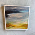 A square framed abstract landscape painting with textured brushstrokes in gray, black, blue, yellow, and orange, displayed on a light gray concrete wall.
