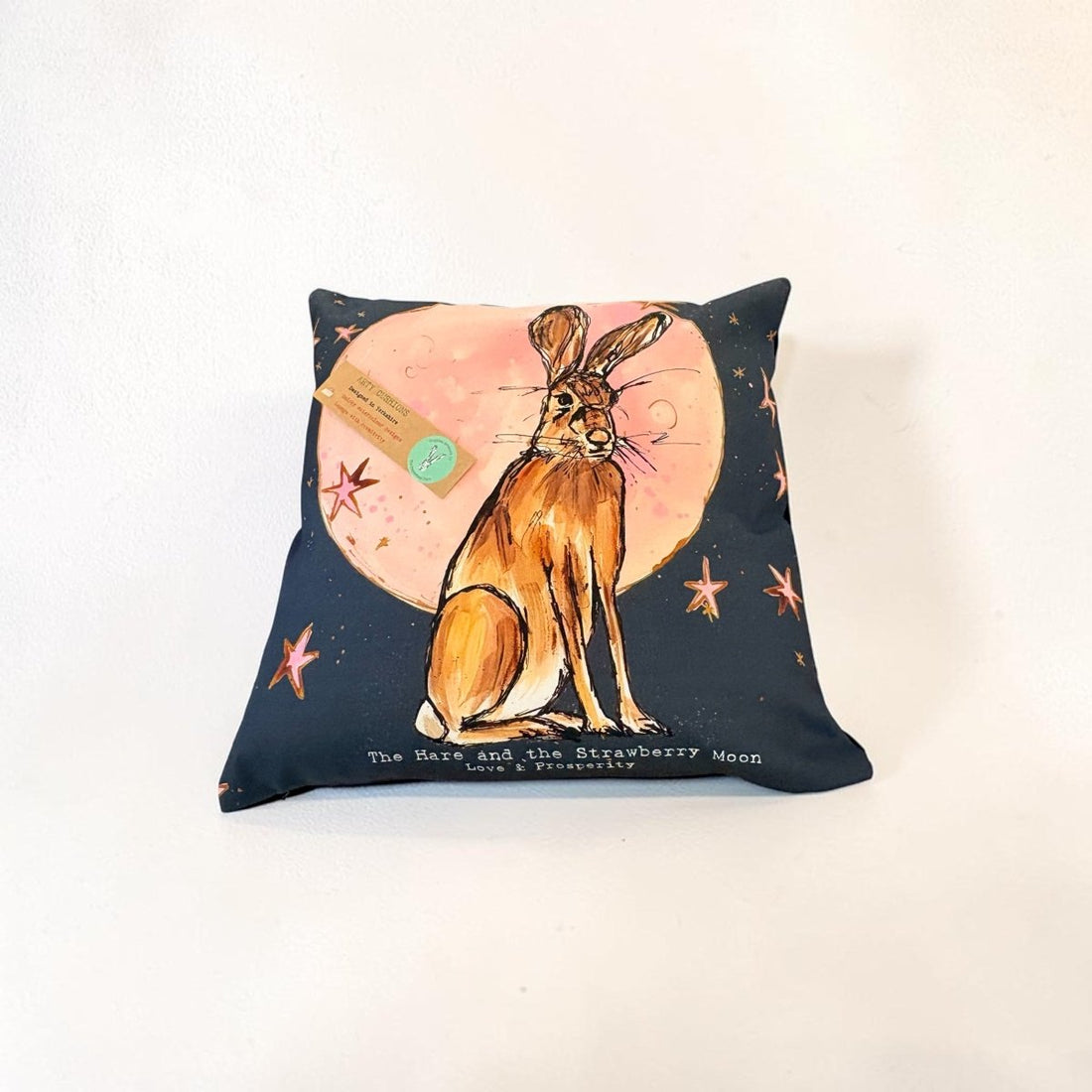 A square decorative pillow featuring a watercolor-style illustration of a brown hare sitting in front of a pink moon, with small orange stars on a dark navy background. A small paper tag is attached to the pillow, and the text &