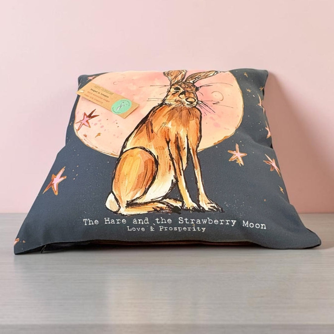 A square decorative pillow on a gray surface against a light pink wall. The pillow features a watercolor-style illustration of a brown hare sitting in front of a pink strawberry moon, with small orange starfish and the text &