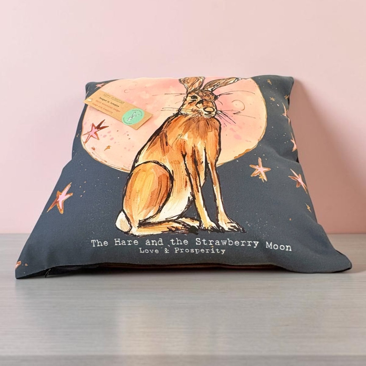 A square decorative pillow on a gray surface against a light pink wall. The pillow features a watercolor-style illustration of a brown hare sitting in front of a pink strawberry moon, with small orange starfish and the text &
