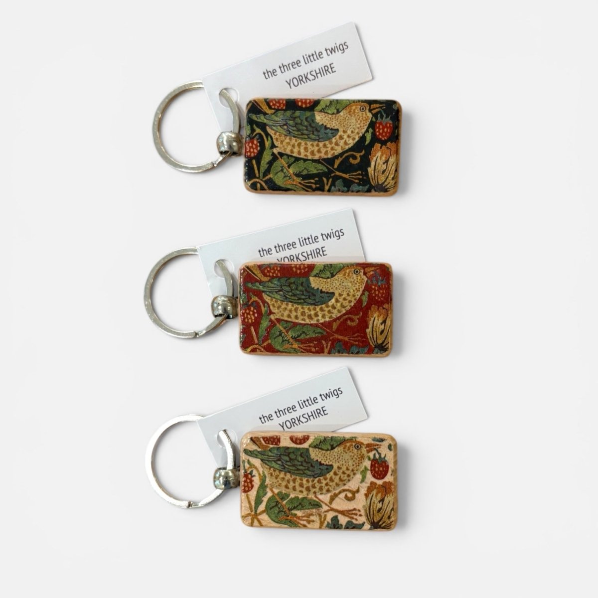 Three rectangular keychains with silver metal key rings, each featuring a different William Morris-style floral and bird pattern in red, green, and beige tones, with white paper tags reading &