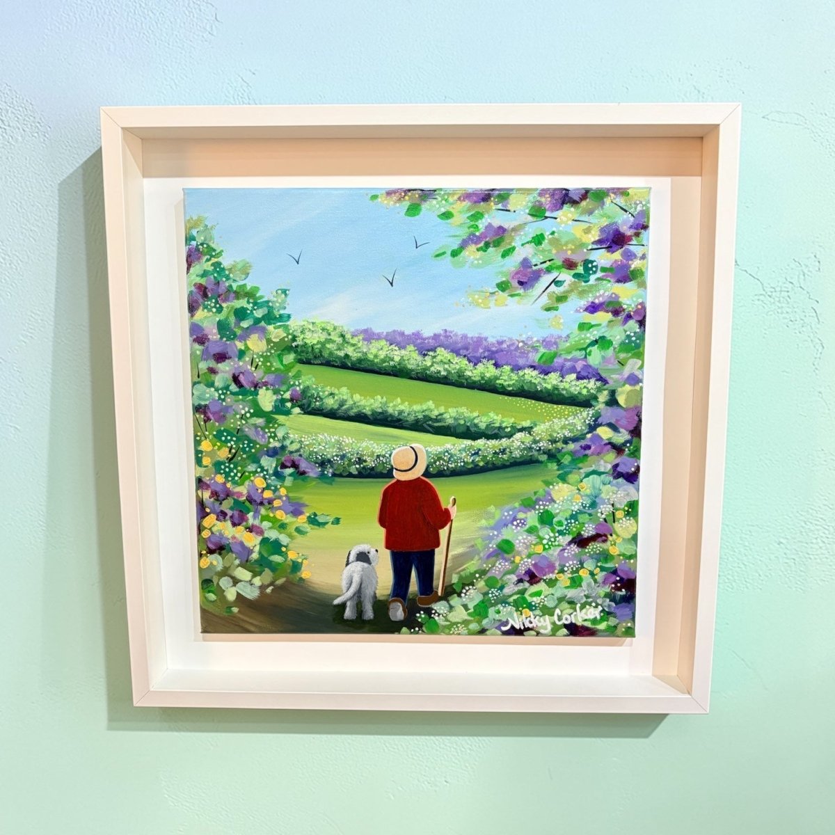 Framed artwork of a person in a garden with flowers and a dog on a light blue wall.