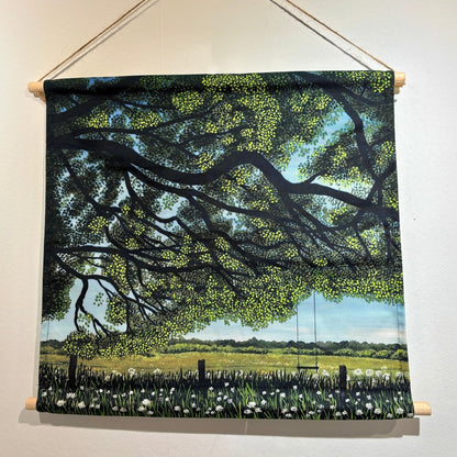 A fabric wall hanging scroll depicting a scenic landscape with a large tree, green field, flowers, and a swing, suspended by twine from wooden dowels on a white wall.