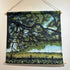A fabric wall hanging scroll depicting a scenic landscape with a large tree, green field, flowers, and a swing, suspended by twine from wooden dowels on a white wall.