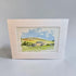 A framed watercolor landscape print featuring rolling green hills, a small house, trees, and a blue sky, displayed against a neutral light gray background.