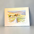 A watercolor landscape art print depicting a rural scene with a winding path, small cottages, trees, and rolling hills, shown with a wide cream mount against a neutral gray background.