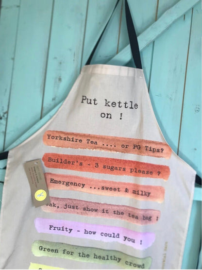 A beige canvas kitchen apron with black handles, featuring humorous tea-themed text printed on it over pastel stripes. The apron is displayed against a light blue wooden plank background.