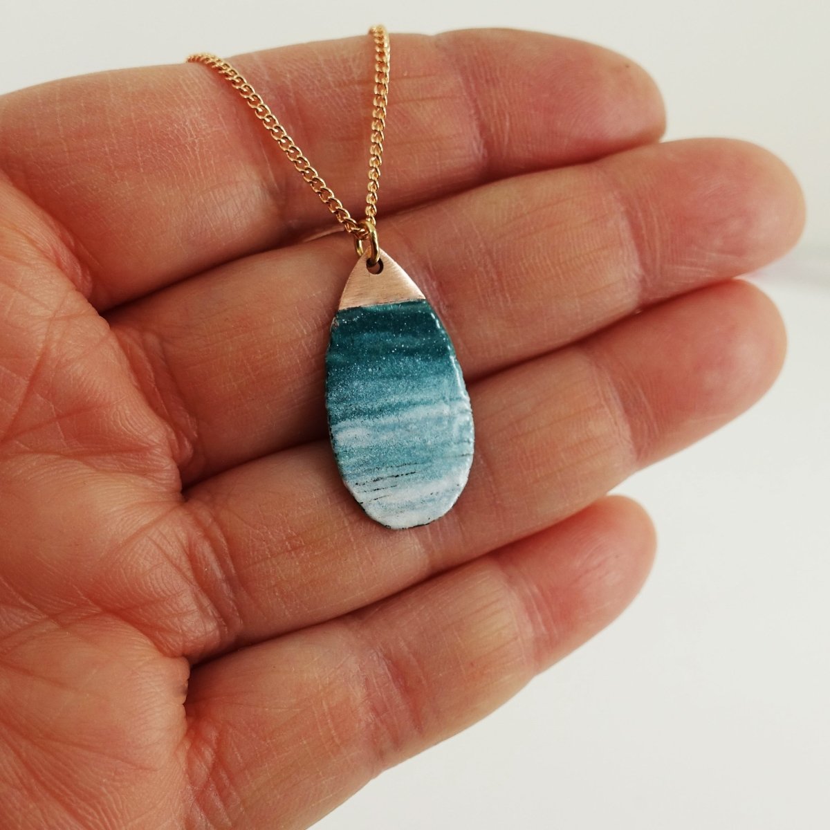 Teal and white marbled teardrop pendant on a gold chain held in a hand against a white background