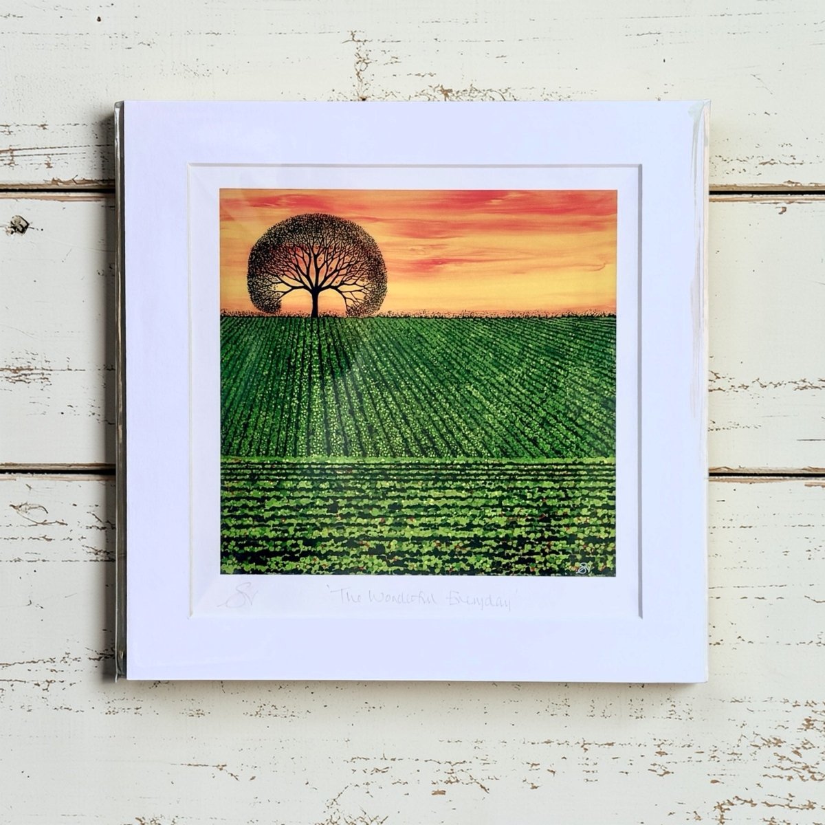 Framed artwork of a tree over a field with a sunset sky on a wooden surface.