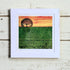 Framed artwork of a tree over a field with a sunset sky on a wooden surface.