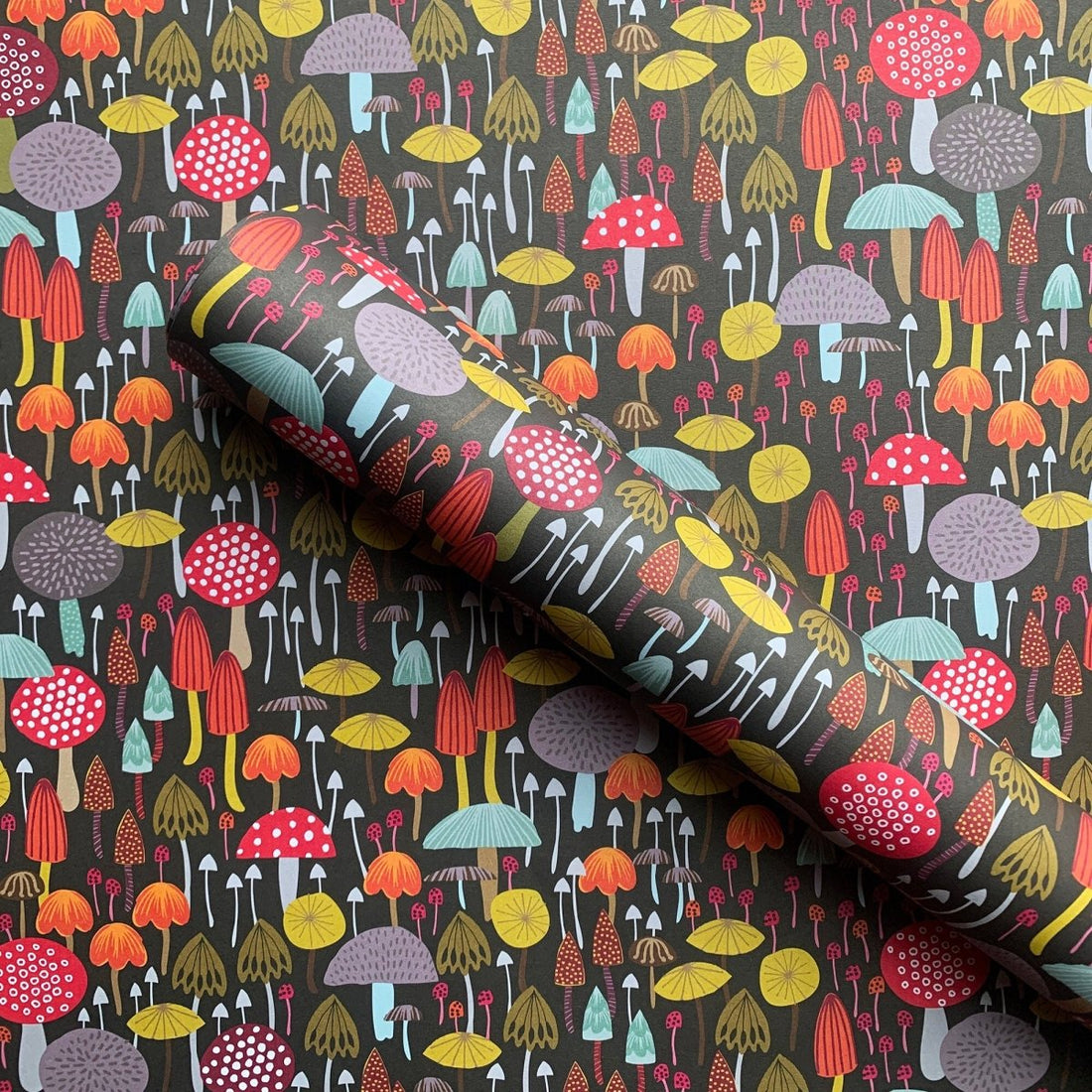 A roll of patterned wallpaper or fabric featuring a dense, whimsical mushroom design on a dark charcoal background. The pattern includes mushrooms in red, orange, yellow, purple, and light blue, with some featuring white polka dots.