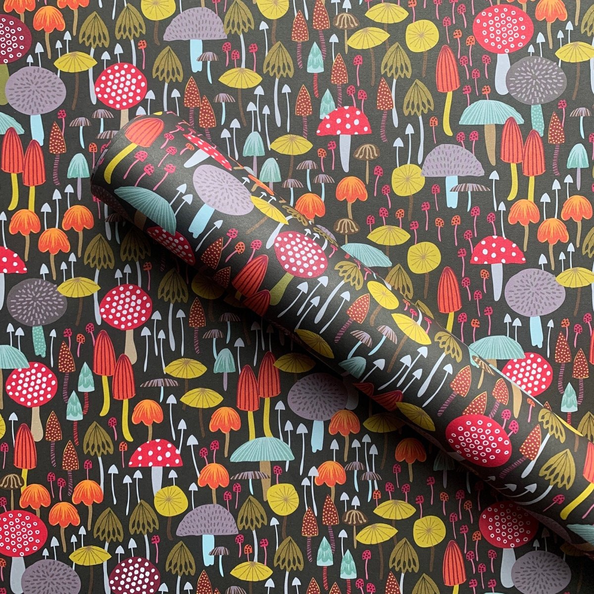 A roll of patterned wallpaper or fabric featuring a dense, whimsical mushroom design on a dark charcoal background. The pattern includes mushrooms in red, orange, yellow, purple, and light blue, with some featuring white polka dots.