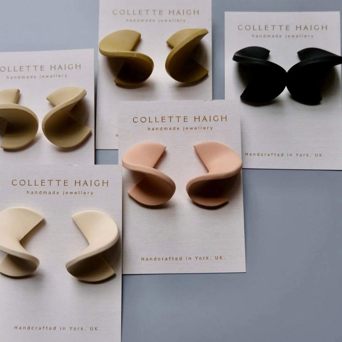 Six pairs of Collette Haigh handmade jewelry earrings in various neutral colors (black, beige, cream, peach, olive, and light pink) displayed on their white branded backing cards against a cool gray background.