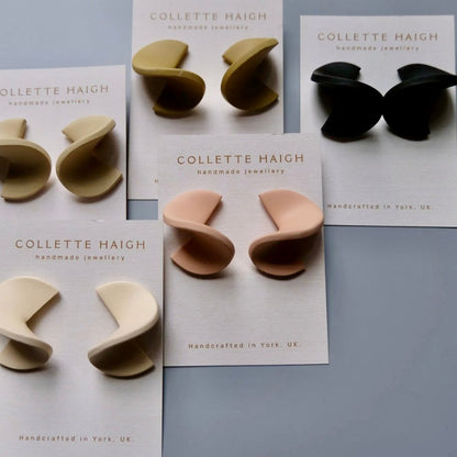 Six pairs of Collette Haigh handmade jewelry earrings in various neutral colors (black, beige, cream, peach, olive, and light pink) displayed on their white branded backing cards against a cool gray background.