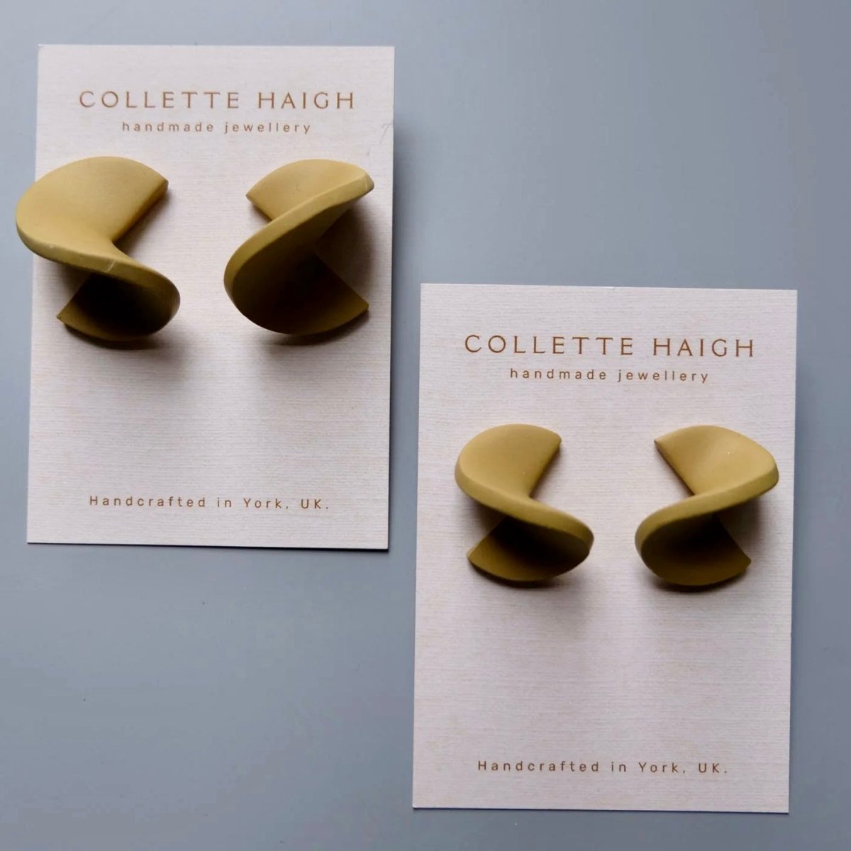 Two pairs of Collette Haigh handmade spiral stud earrings in a muted olive-gold tone, mounted on white display cards against a light gray background.