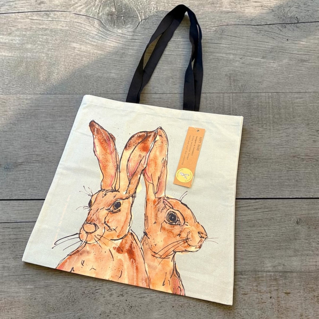 A cream canvas tote bag with black shoulder straps, featuring a watercolor-style illustration of two brown hares. A small orange paper tag is attached to the bag, which is photographed from above on a gray wooden plank surface.