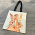 A cream canvas tote bag with black shoulder straps, featuring a watercolor-style illustration of two brown hares. A small orange paper tag is attached to the bag, which is photographed from above on a gray wooden plank surface.