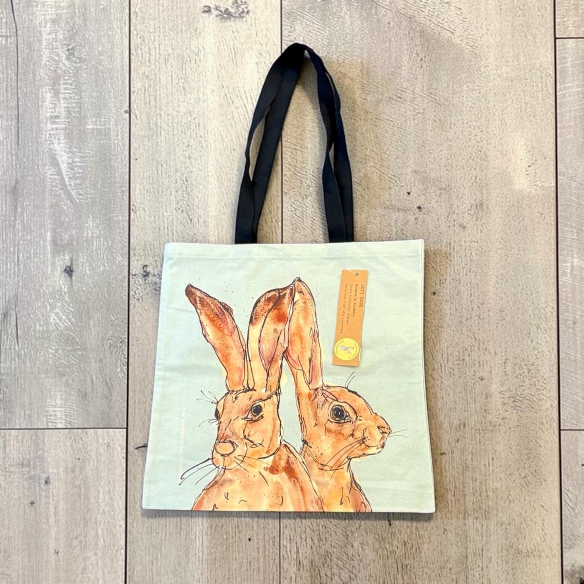 A canvas tote bag with black shoulder straps, featuring a watercolor-style illustration of two brown hares on a pale mint background. The bag is laid flat on a light gray wood plank floor, with a small yellow paper tag attached.