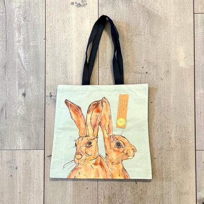 A canvas tote bag with black shoulder straps, featuring a watercolor-style illustration of two brown hares on a pale mint background. The bag is laid flat on a light gray wood plank floor, with a small yellow paper tag attached.