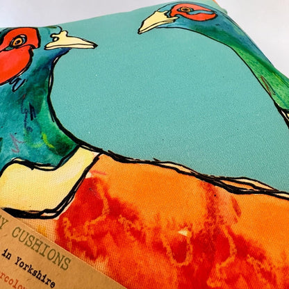 A close-up of a decorative throw pillow featuring a colorful printed design of pheasants on a turquoise background, with part of a brown paper label visible at the bottom left.
