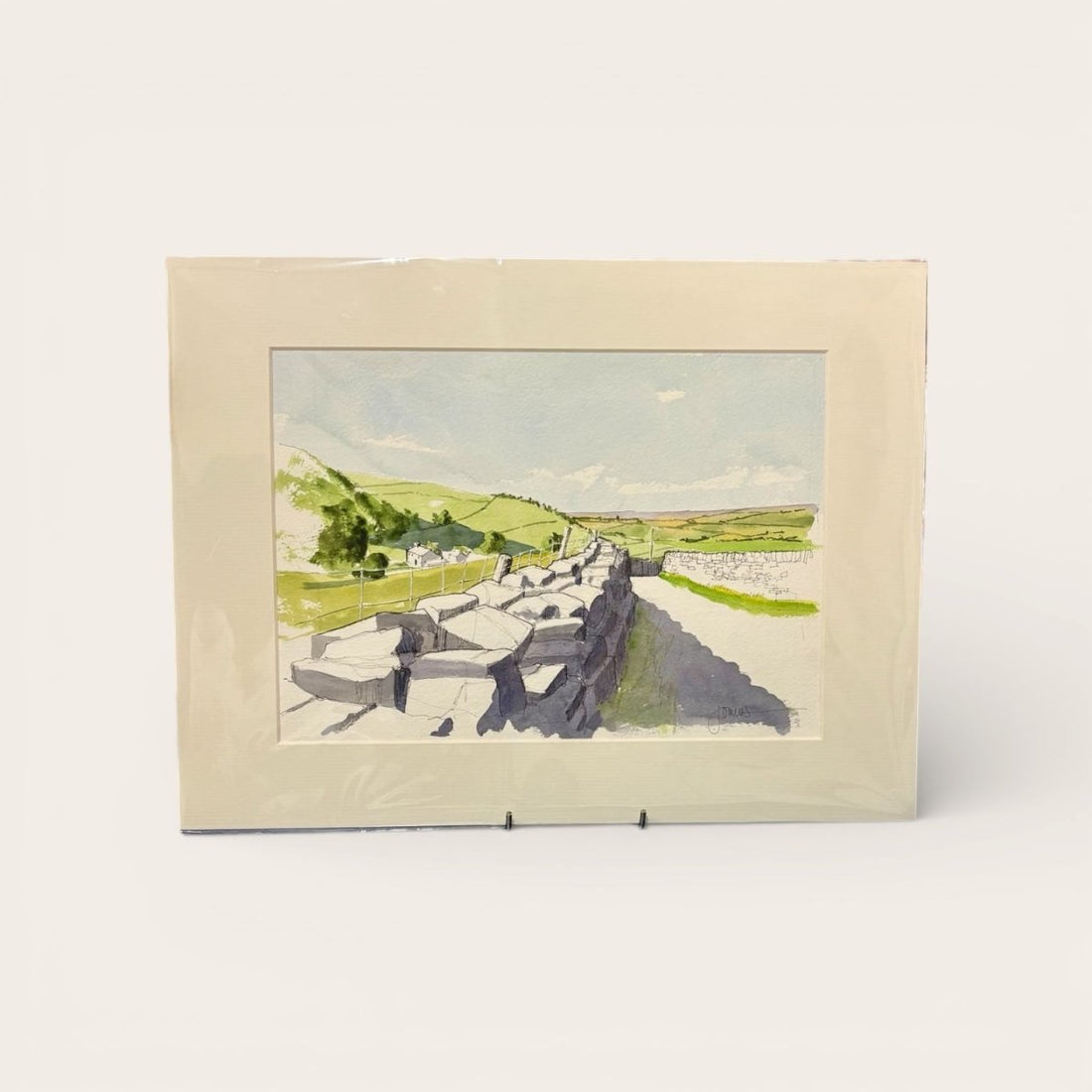Framed watercolor landscape painting of a stone wall and green fields.