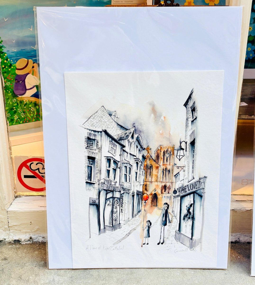A matted art print depicting a street scene with old buildings and a small figure in the foreground, photographed leaning against a shop window. The print is wrapped in protective plastic, and surrounding storefront elements are partially visible.